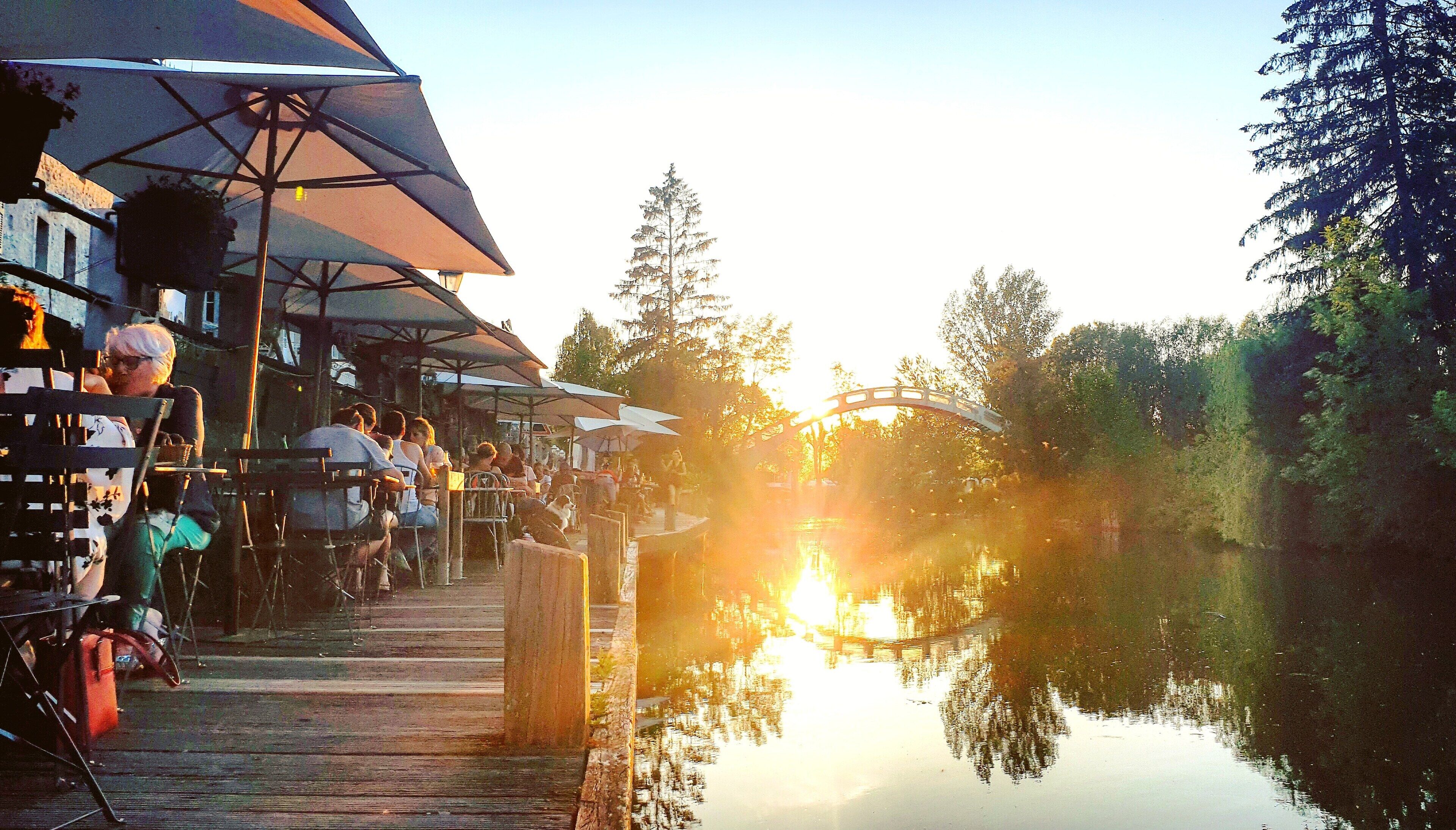 Un espace de restauration au bord de la rivière, avec des tables à l’extérieur sous des parasols, une terrasse en bois et le reflet des arbres dans l’eau.
