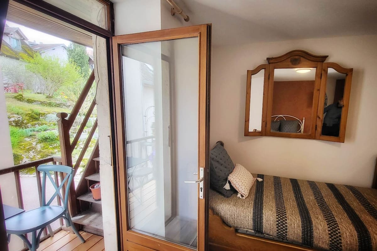 A bedroom with a bed, a chair, a wooden-framed mirror, and a sliding glass door leading to a balcony.