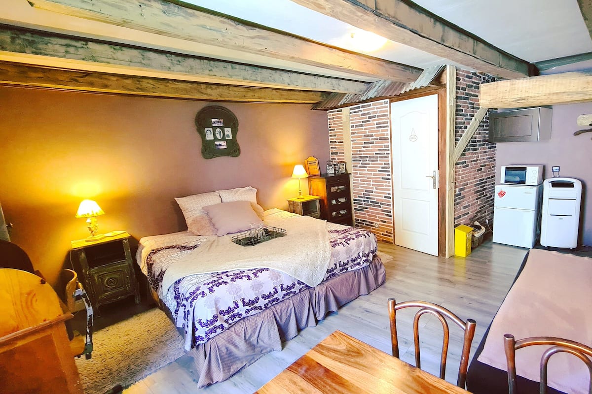 A cozy bedroom with a bed, bedside table, lamp, dresser, and a small kitchenette area with a fridge and microwave.