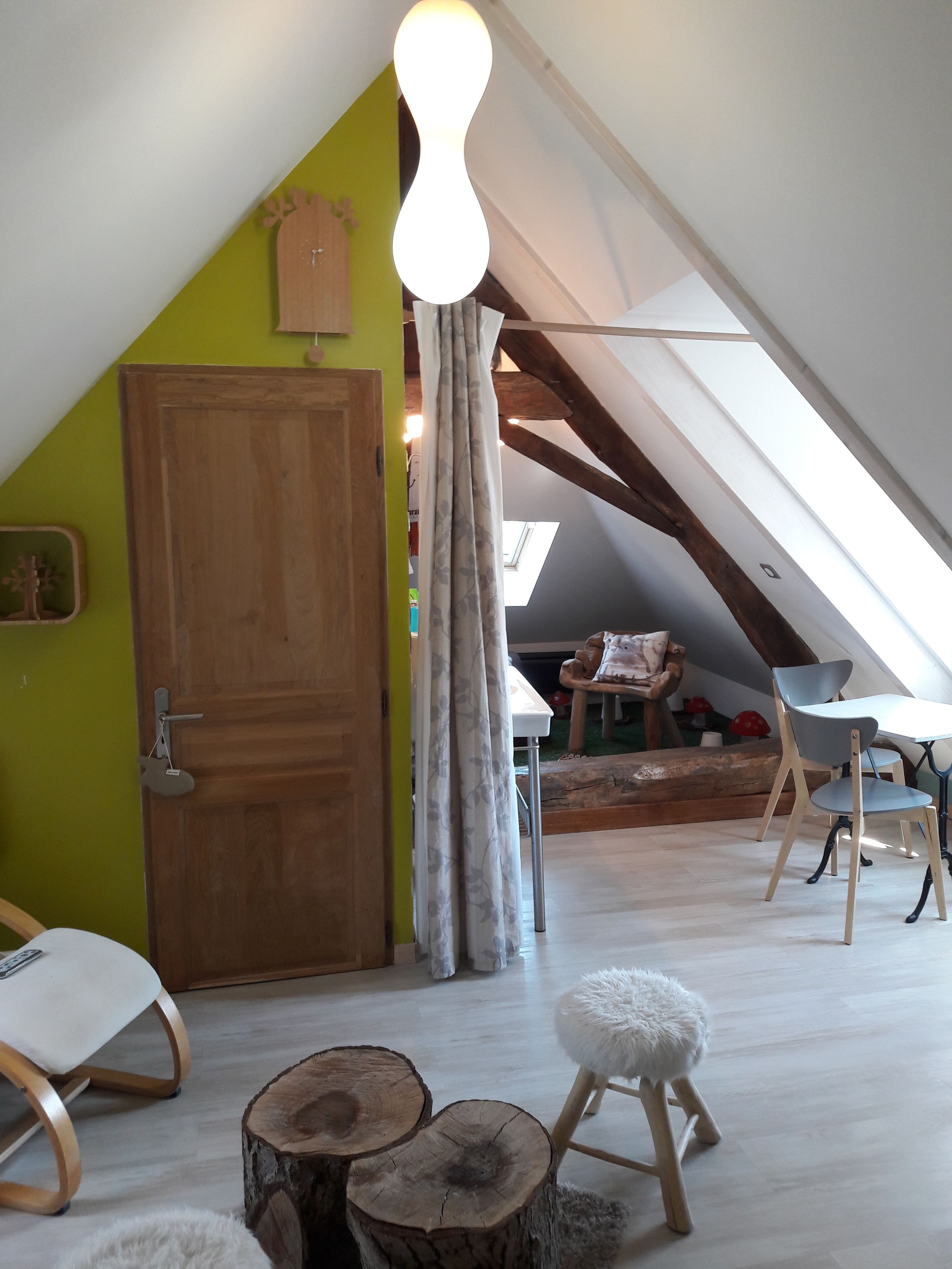 Une pièce mansardée chaleureuse, avec un plafond en pente, une porte en bois et un puits de lumière. On y trouve deux tabourets en bois et un pouf blanc moelleux.