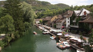 Eine Flussstadt mit vor Anker liegenden Booten, mit traditioneller Architektur versehenen Gebäuden und einem bewaldeten Hügel im Hintergrund.