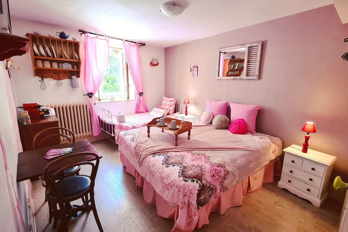 A cozy bedroom with a large bed, a small desk, and a chair. There is a radiator, a window with curtains, and a bedside table with a lamp.