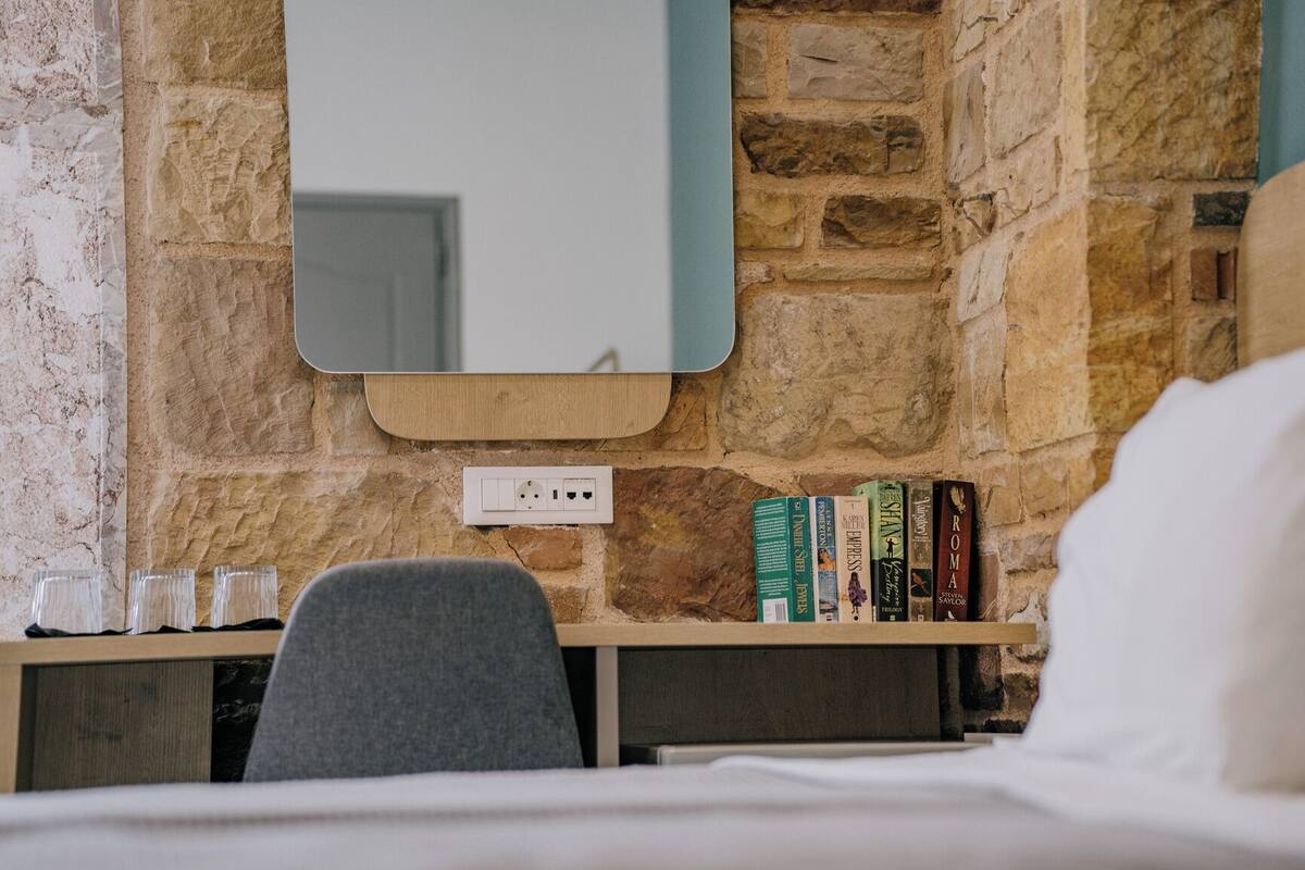 A hotel room with a stone wall, a wooden desk, a mirror, and a bookshelf with books.