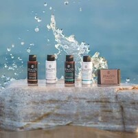 Cuatro botellas de productos Zealots of Nature y una caja de Jabón Mandarin Zealots of Nature sobre una superficie de piedra con salpicaduras de agua al fondo.