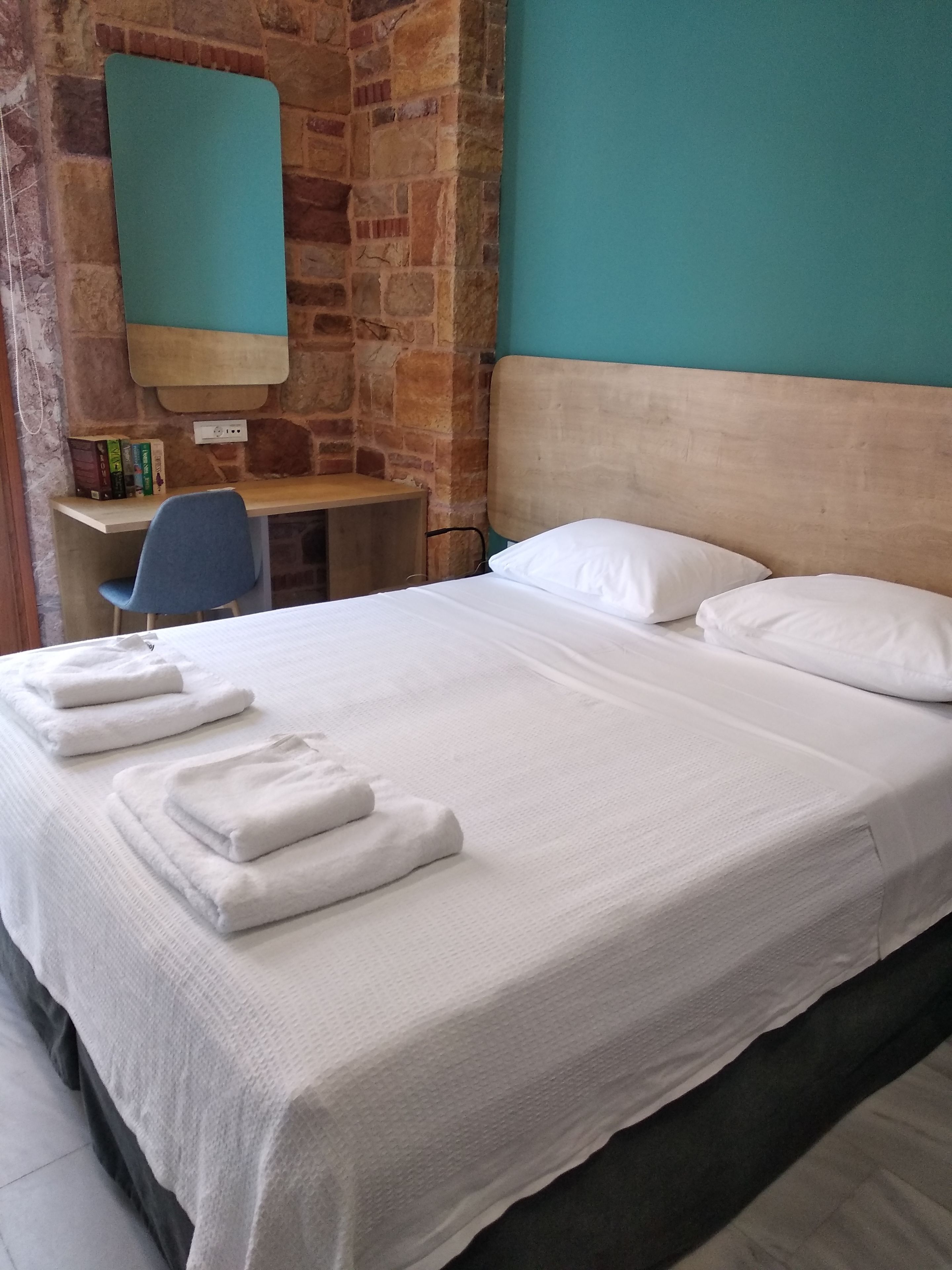 Een net opgemaakt bed met witte lakens en handdoeken, een houten bureau met een stoel, en een stenen muur.
