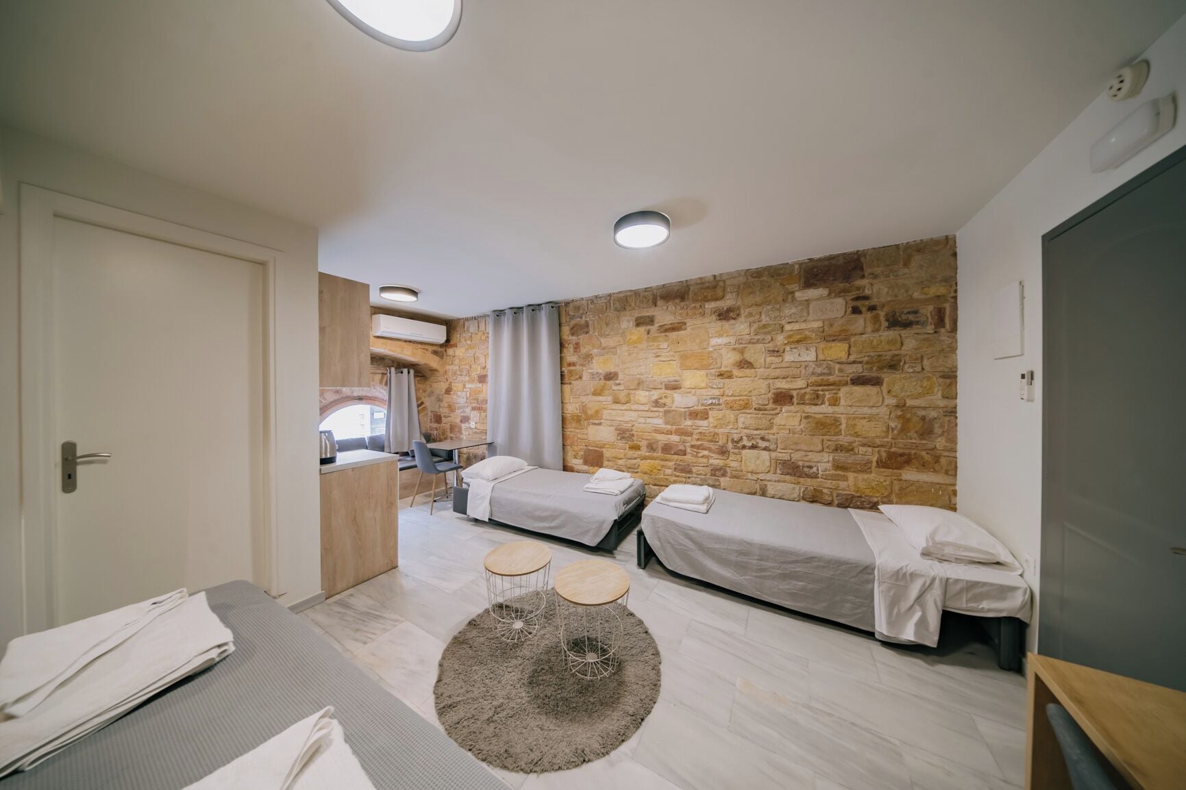 Een compacte hotelkamer met een stenen muur, een kleine kitchenette, twee eenpersoonsbedden, een ronde tafel met stoelen en een groot raam.