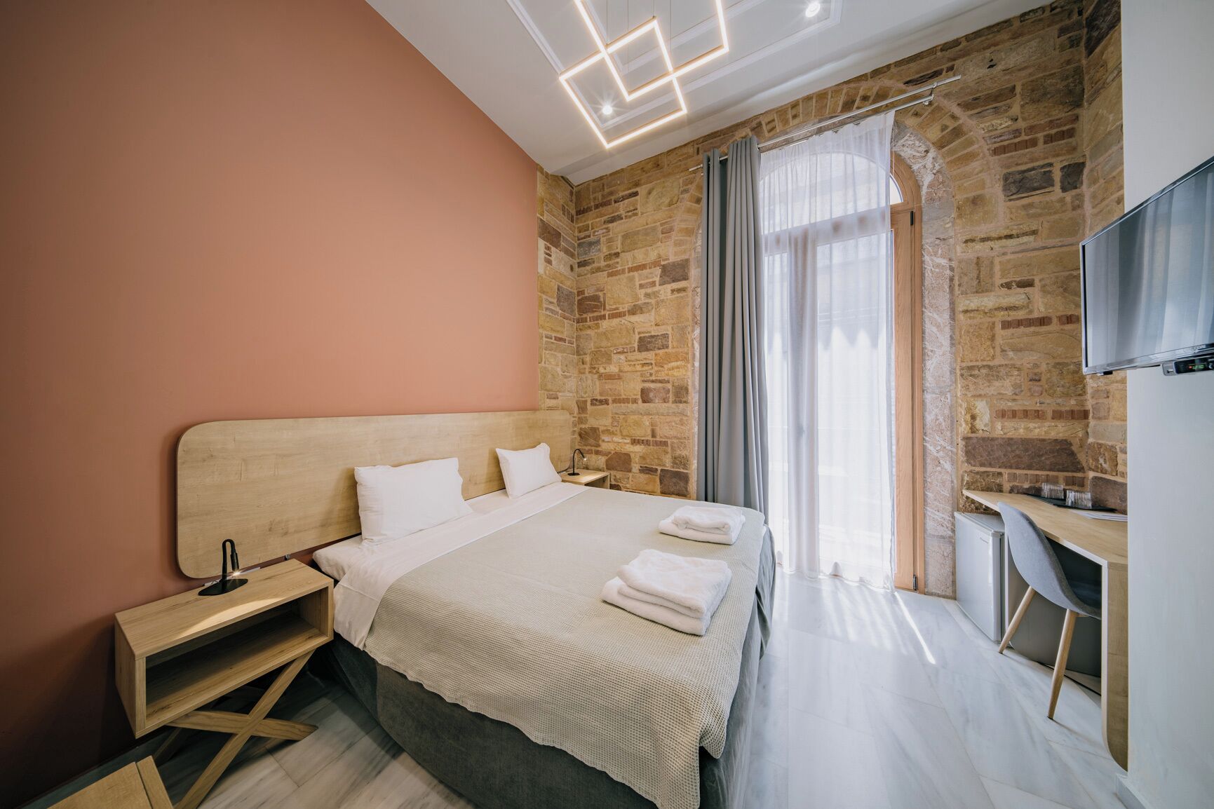 A modern hotel room with a stone wall, a wooden bed, a desk, and a TV.