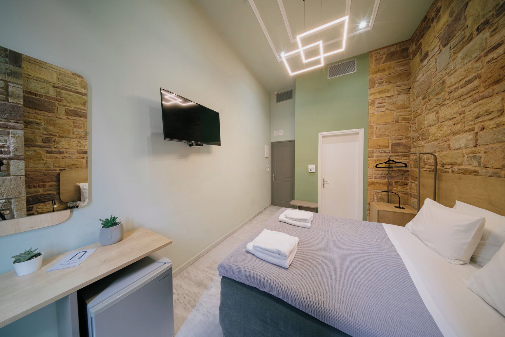 A modern bedroom with a stone wall, a flat-screen TV, a bedside table with a plant, and a neatly made bed.
