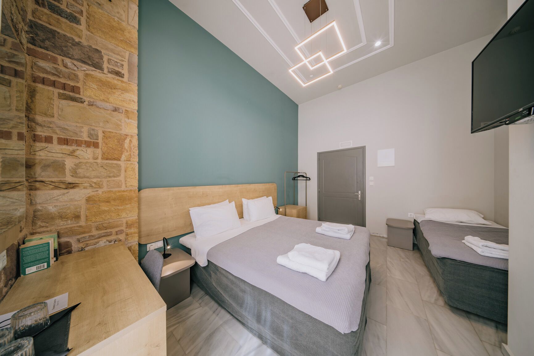 Habitación de hotel con pared de piedra, una cama grande con cabecera, una cama más pequeña, un escritorio con lámpara y un televisor montado en la pared.