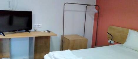 Habitación de hotel con cama, escritorio con televisor, enchufe y estante fijado a la pared.