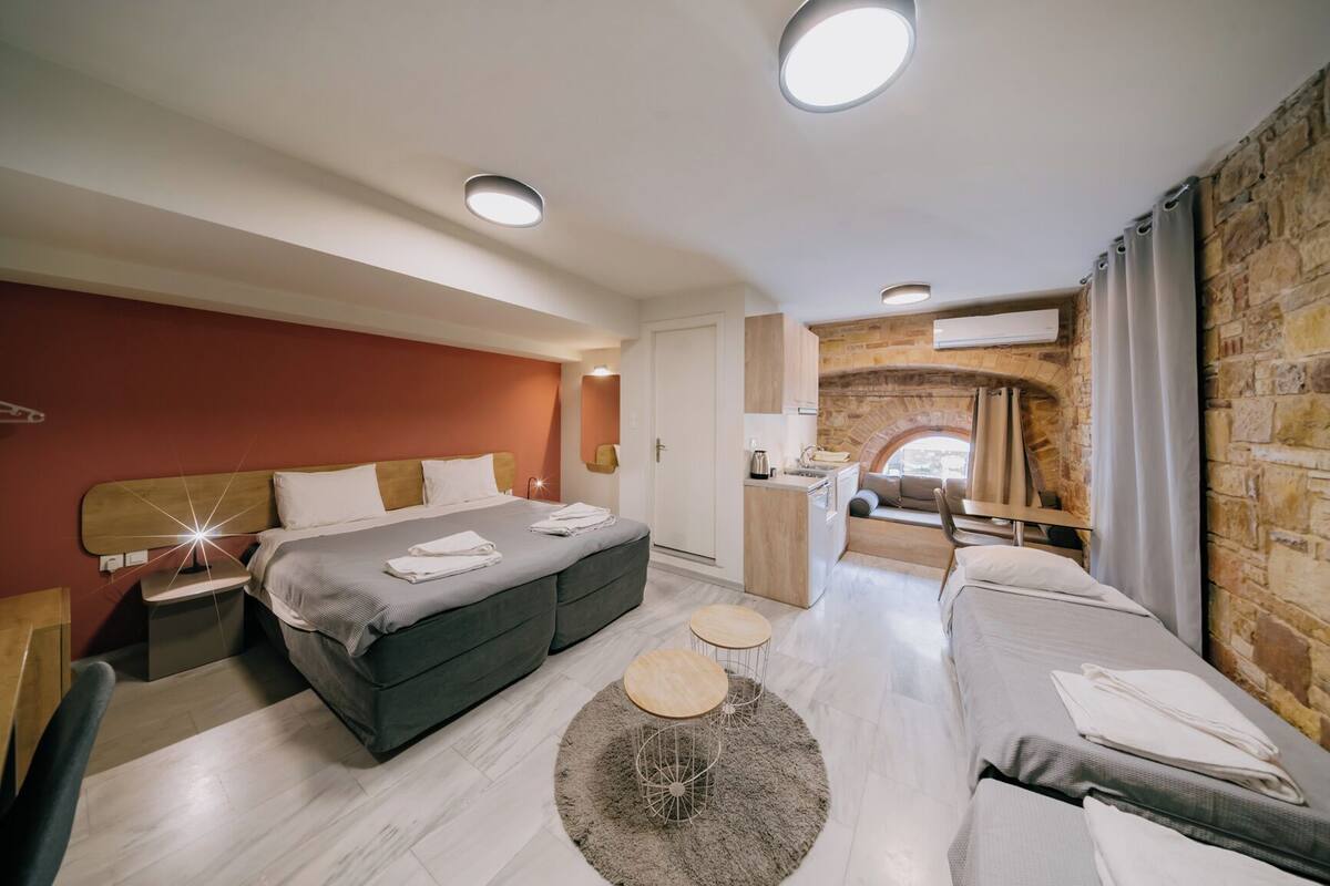 A modern hotel room with a large bed, a sofa, a small kitchen area, and a stone wall.