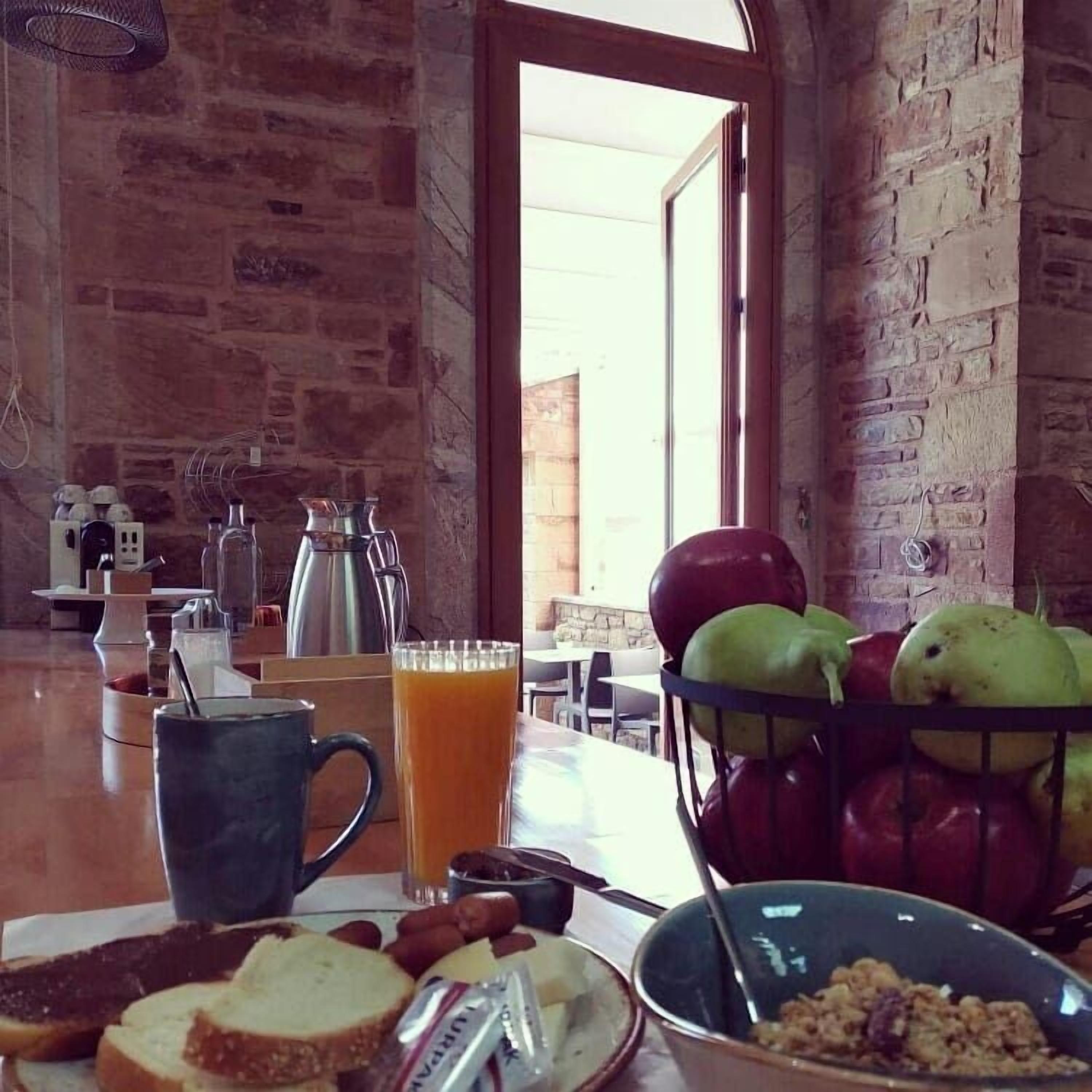 A breakfast table with bread, butter, jam, a bowl of cereal, a glass of orange juice, and a fruit basket.