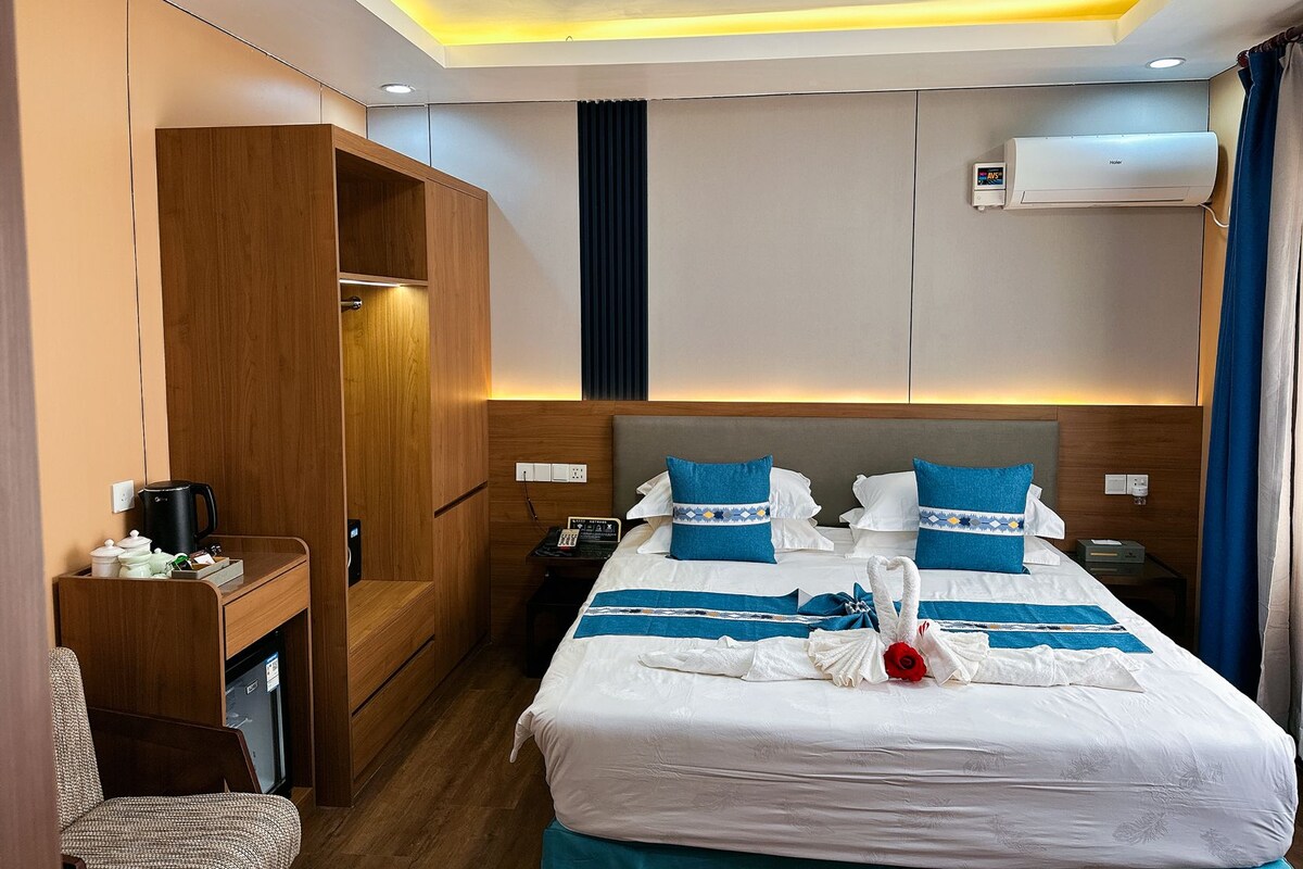 A hotel room with a bed, bedside tables, a desk, a wardrobe, and an air conditioning unit.