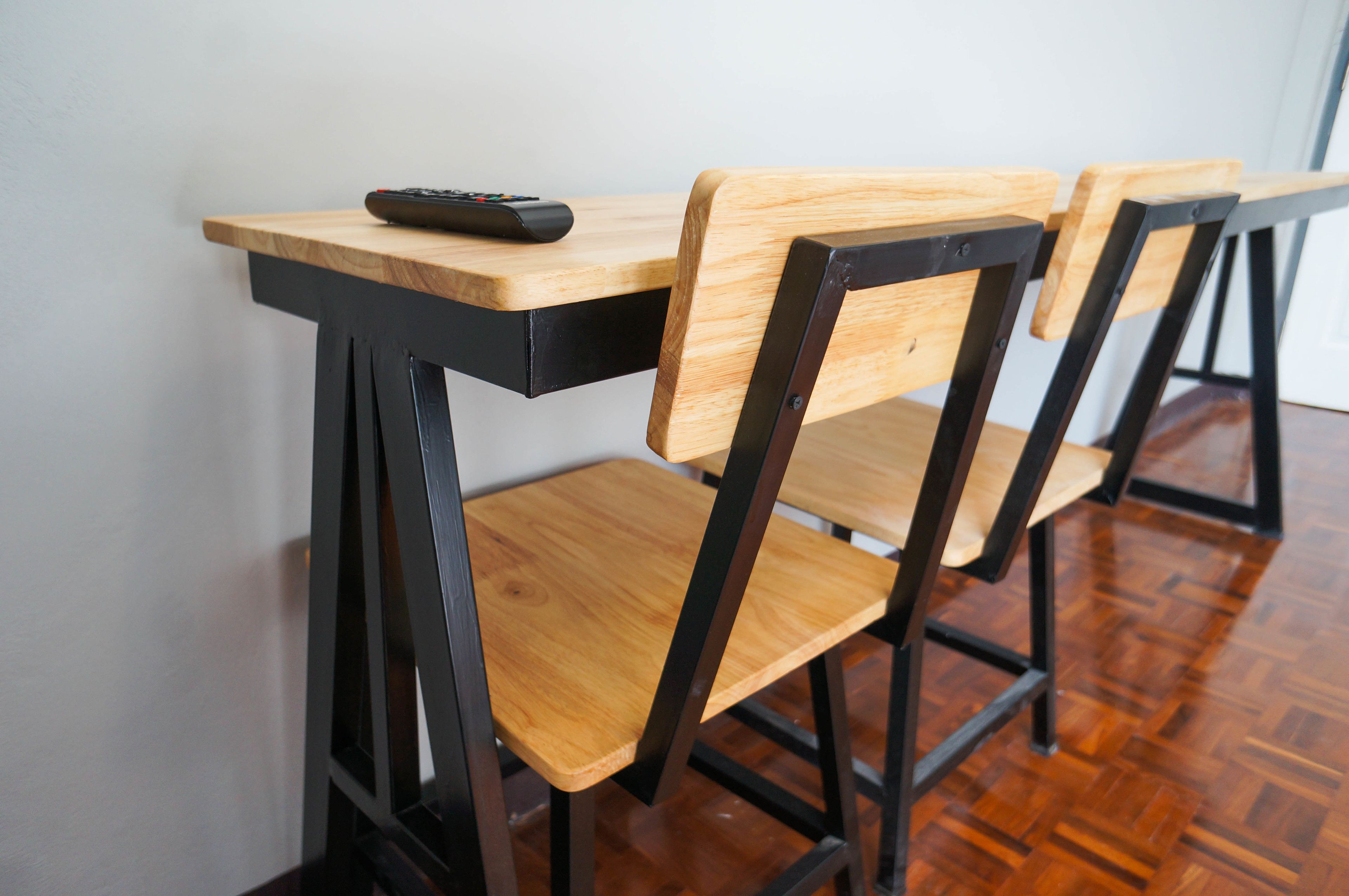 A wooden table with a remote control on it, accompanied by two chairs with black metal frames and wooden seats.