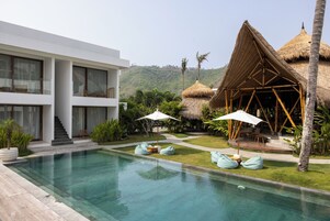 Un resort moderno con piscina, una estructura de techo de paja y un edificio blanco con balcones.