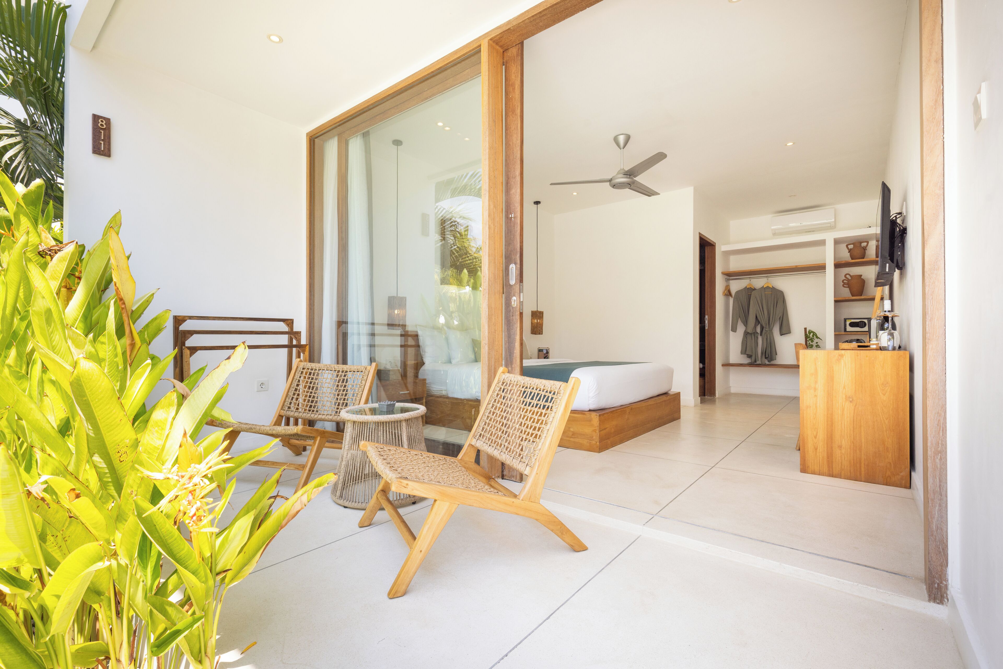 A modern room with a large sliding glass door leading to a balcony, a bed with a wooden headboard, a small seating area with wicker chairs, and a wooden desk with a television.