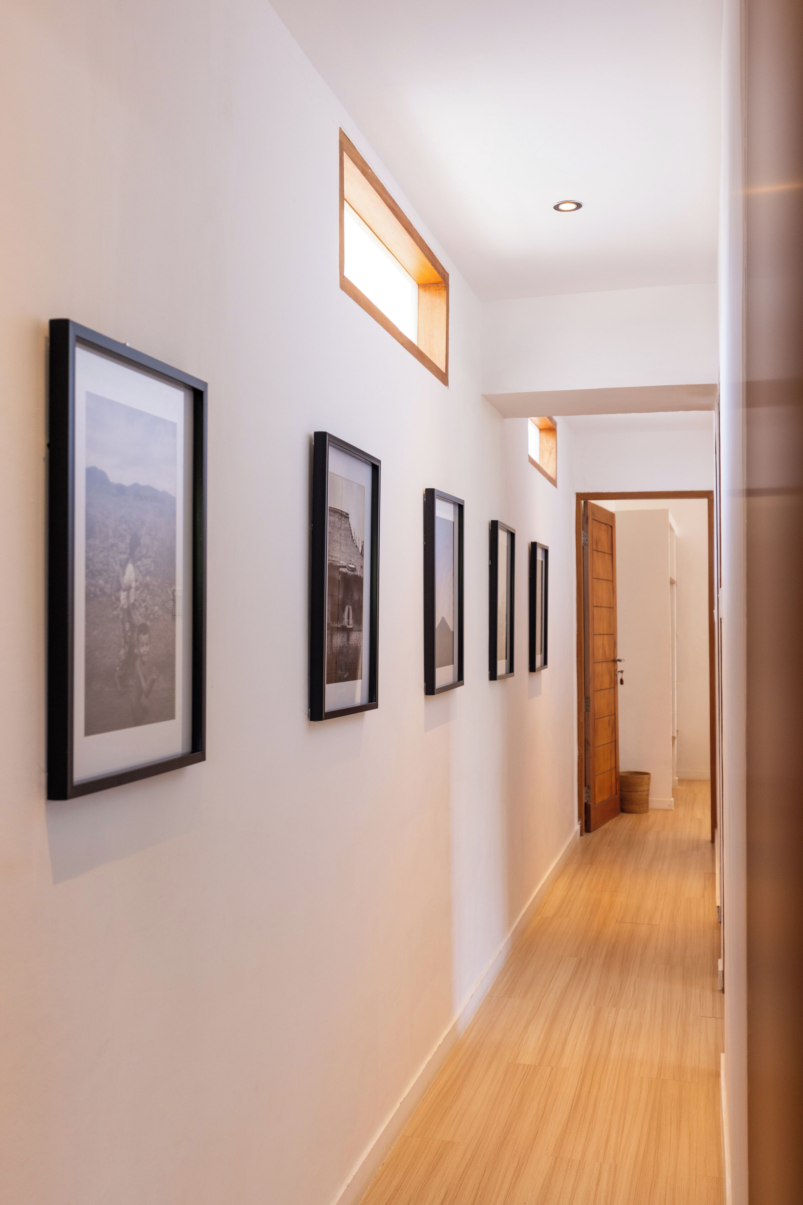 A hallway with framed pictures on the wall, a wooden floor, and a door at the end.