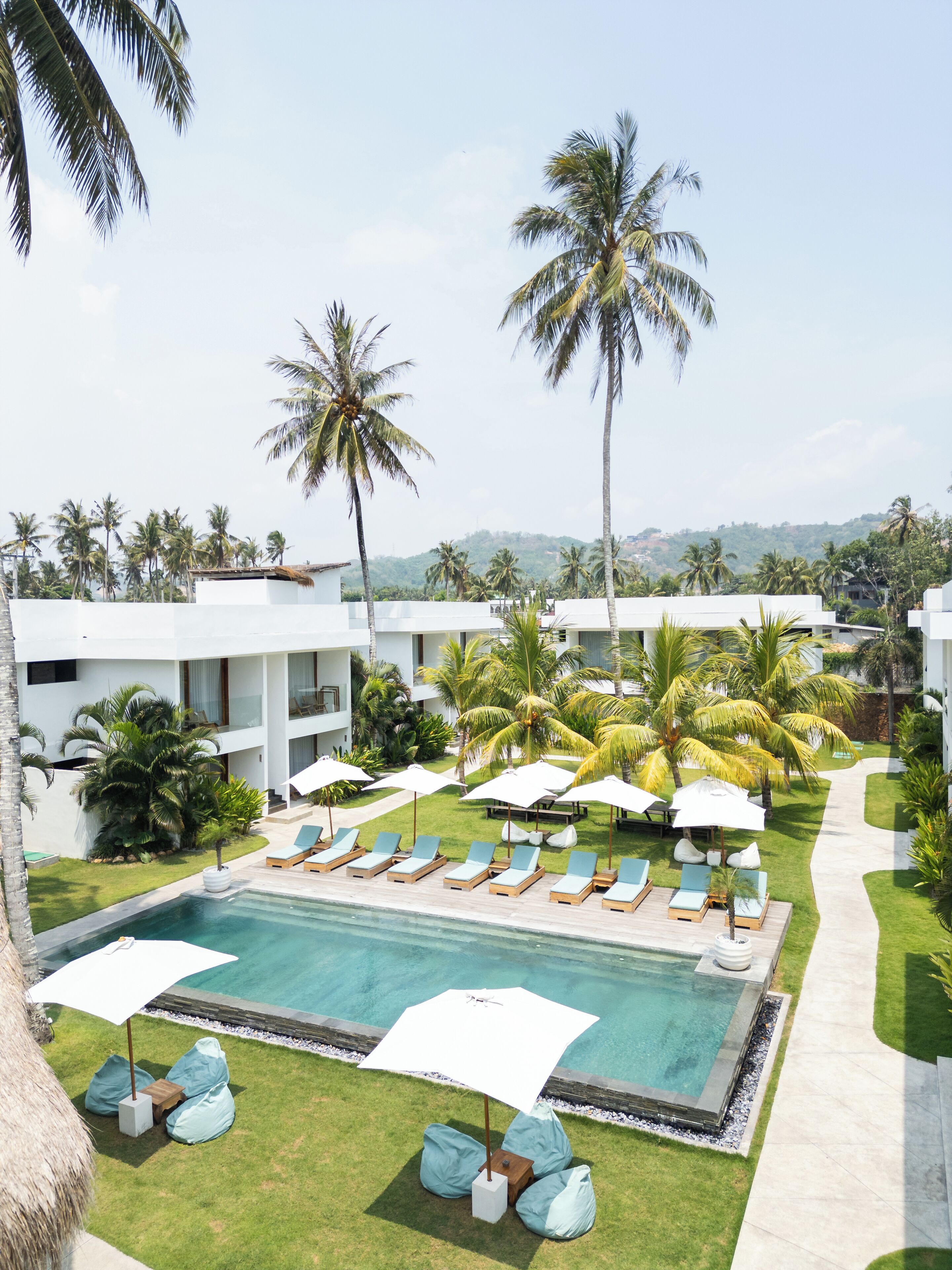 A pool area with lounge chairs and umbrellas, surrounded by palm trees and a view of a hilly landscape.