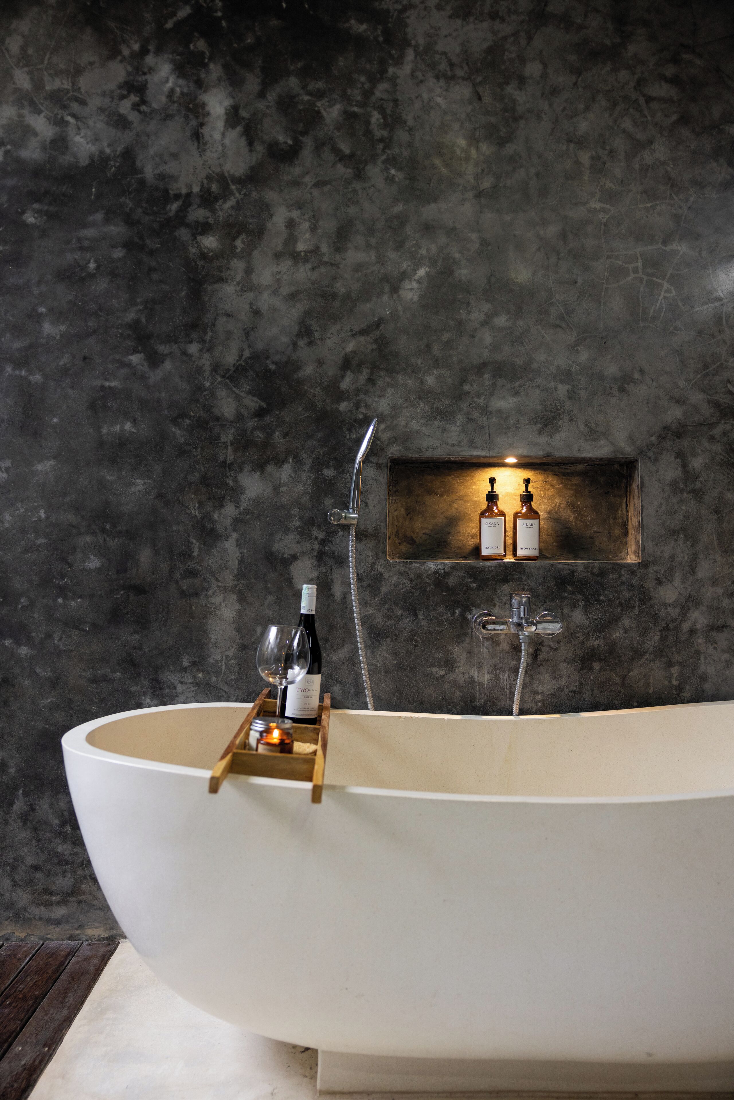 A modern bathroom with a freestanding bathtub, a wooden tray holding a bottle of wine and a glass, and two bottles on a shelf above the tub.