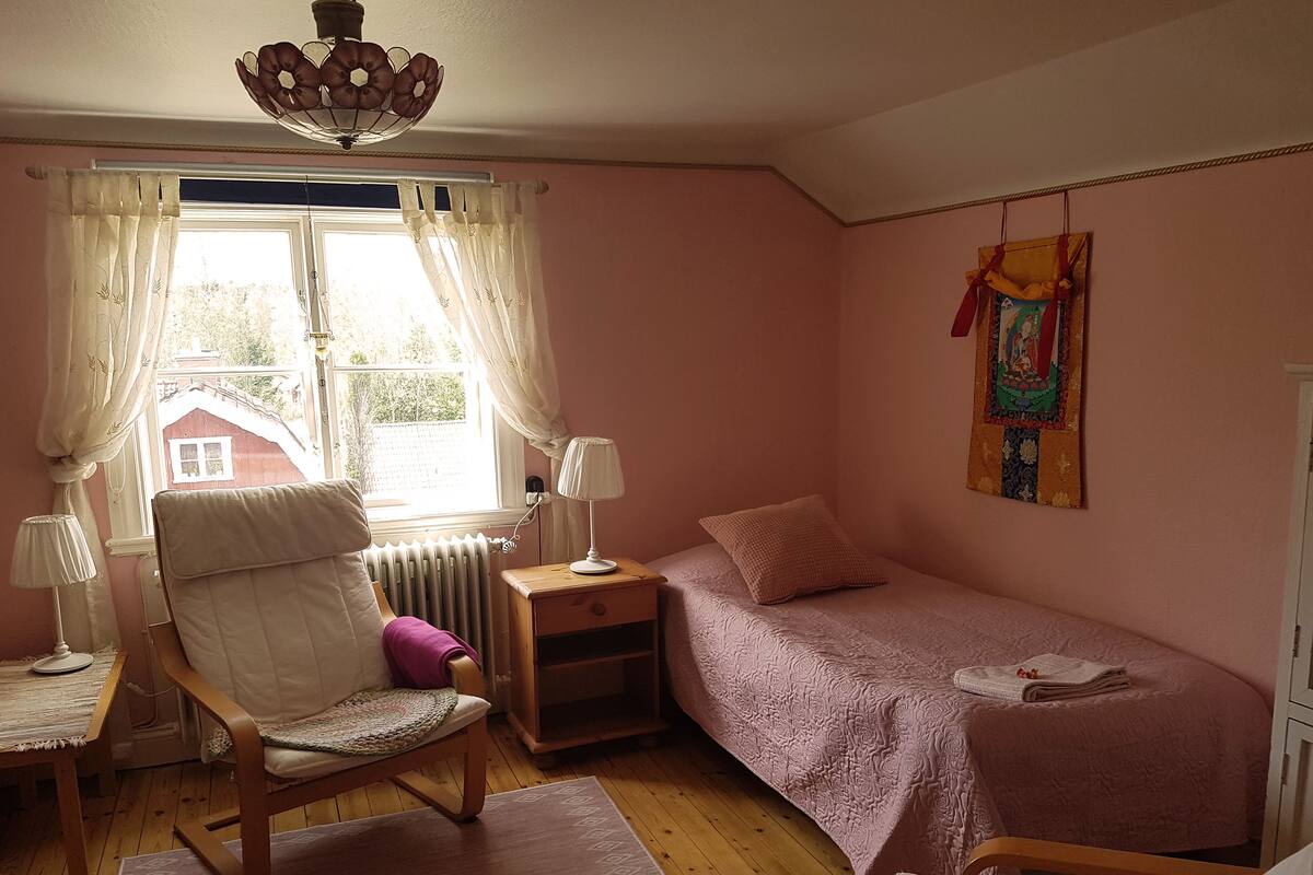 A bedroom with a single bed, a chair, a nightstand, a lamp, a radiator, and a window with curtains.