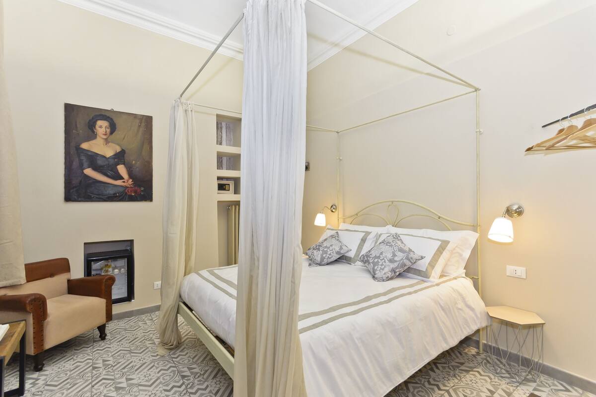 Une chambre à coucher avec un lit à baldaquin, une table de chevet, une chaise, un tableau au mur et une étagère remplie de livres.