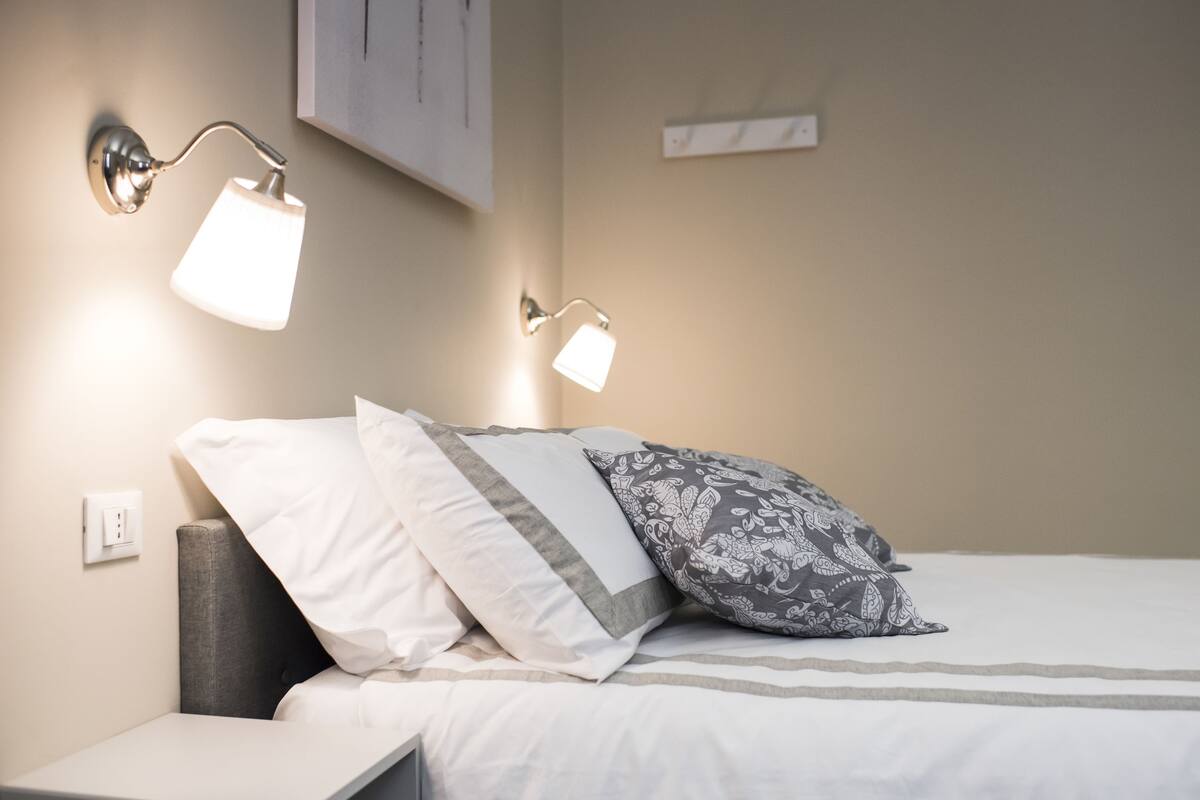 Un lit bien fait, avec une literie blanche et grise, deux lampes de lecture fixées au mur et une petite table de chevet.