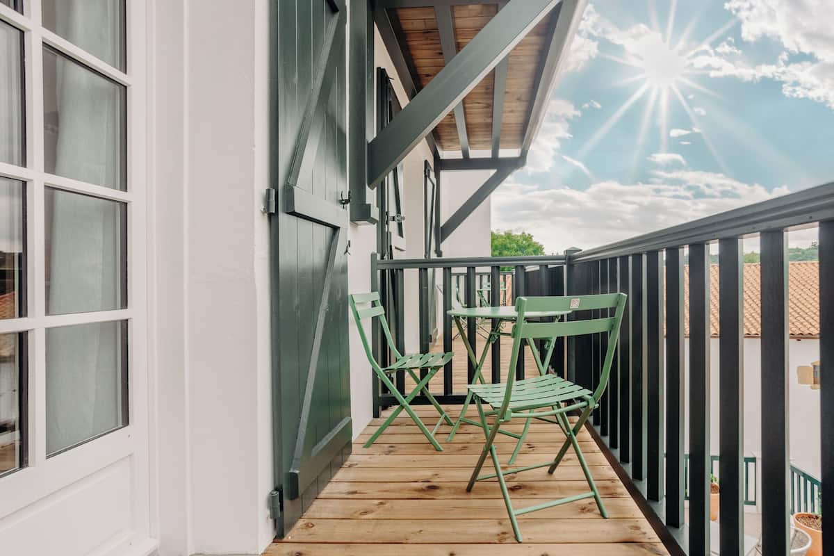 A wooden deck with green chairs and a table, a white building with a black railing, and a clear sky with the sun visible.