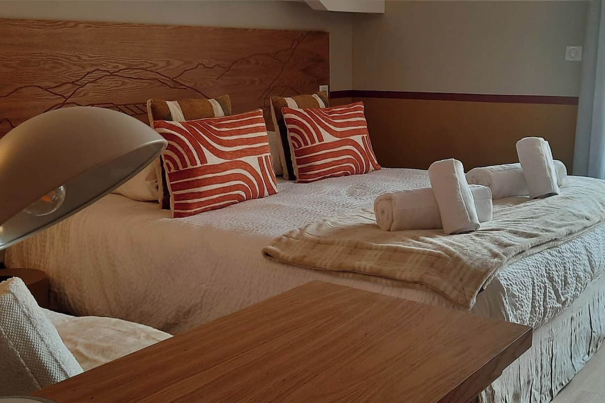 A hotel room with a large bed, a wooden headboard, a desk with a lamp, and a view of the outside through a window.