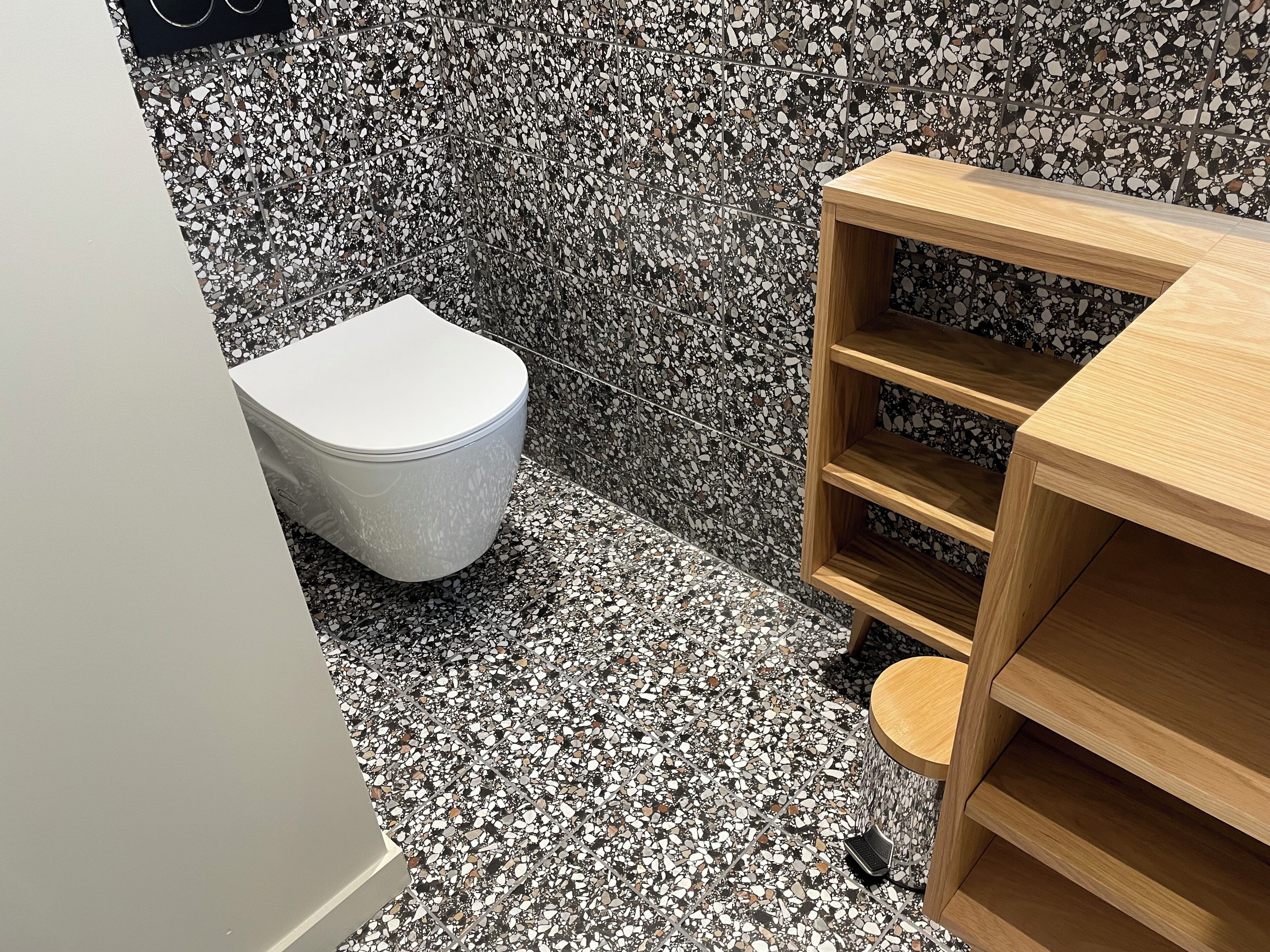 A bathroom with a white toilet, a wooden shelf unit, and a terrazzo floor.