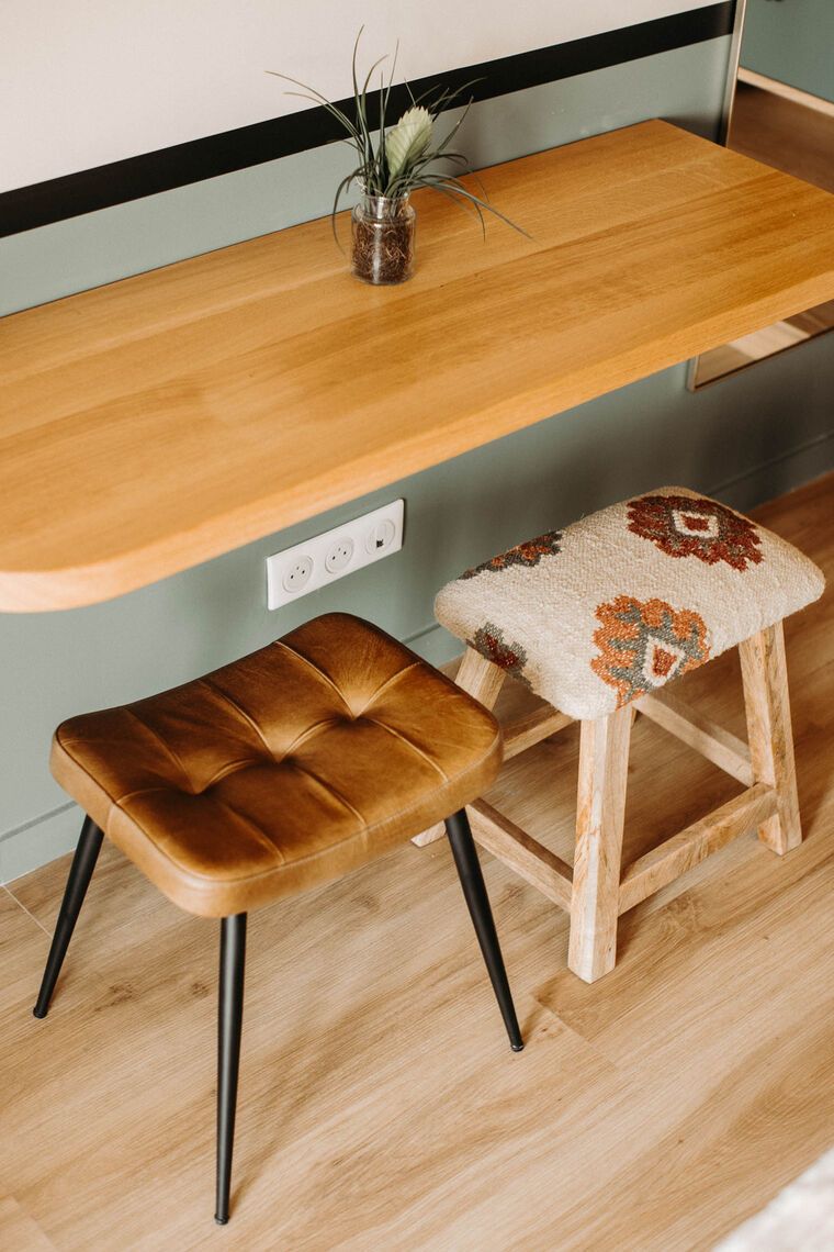 Un bureau en bois sur lequel est posé un vase de plantes, une chaise en cuir marron et un tabouret avec un coussin à motifs.