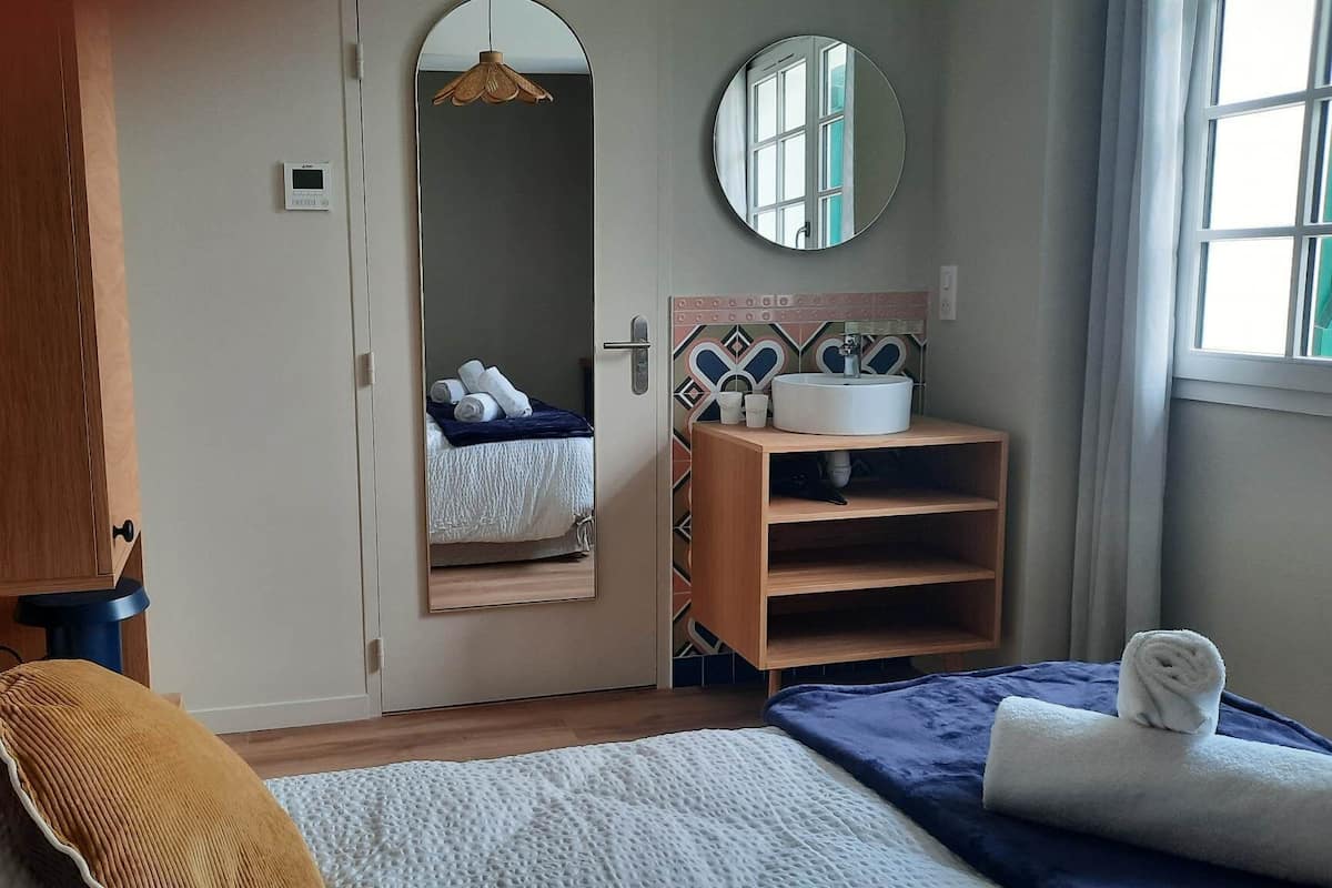 A bedroom with a bed, a bedside table with a mirror, a wooden wardrobe, and a bathroom with a sink and mirror.
