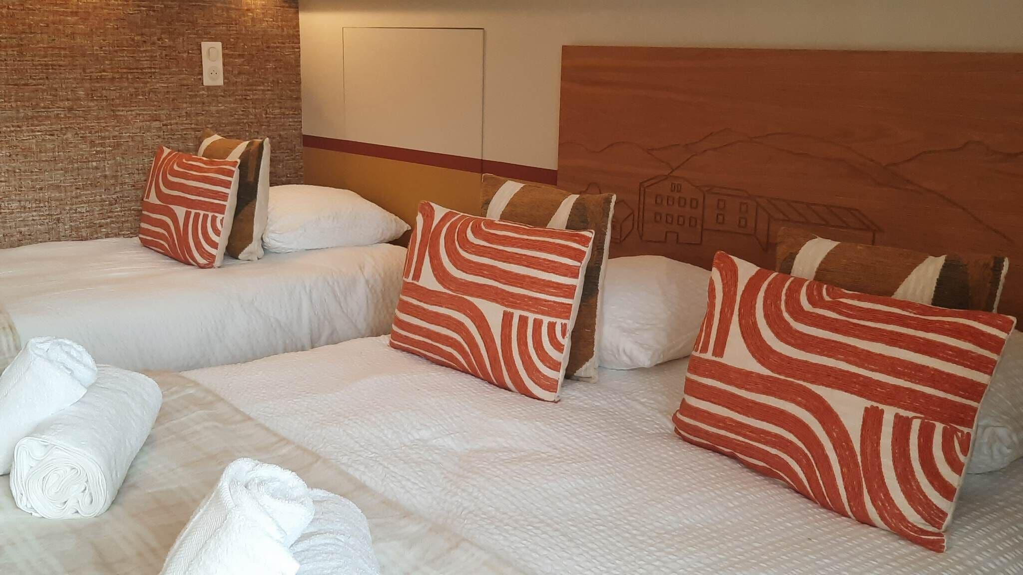 Une chambre d’hôtel avec deux lits, chacun doté de draps blancs et d’oreillers à rayures. On y trouve un mur en briques et une tête de lit en bois ornée d’une fresque.