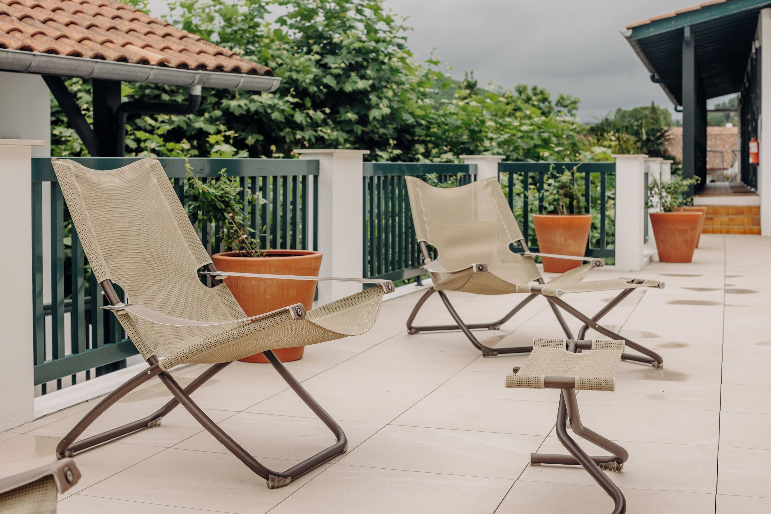 Two lounge chairs with metal frames and mesh seats, positioned on a tiled patio with potted plants and a green fence in the background.