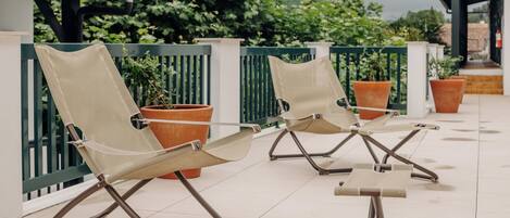 Two lounge chairs with metal frames and mesh seats, positioned on a tiled patio with potted plants and a green fence in the background.