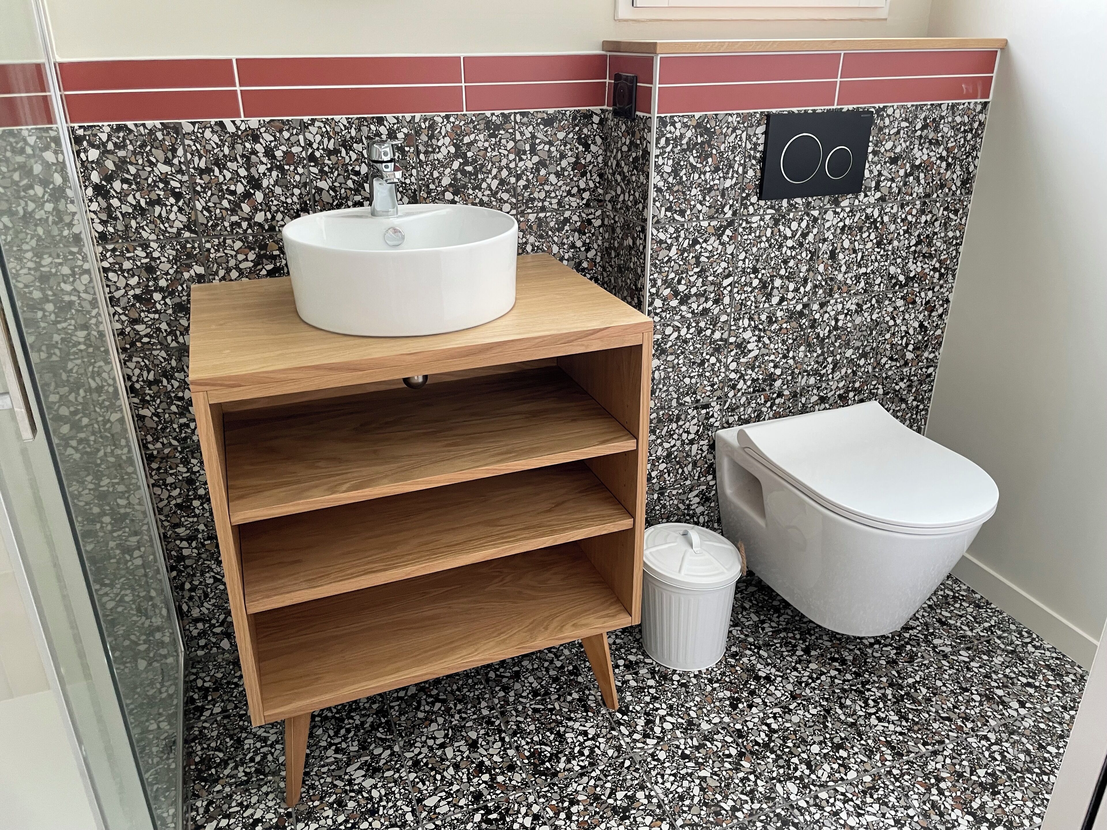Un baño con un lavamanos blanco, repisas de madera, un inodoro y un cubo de basura.