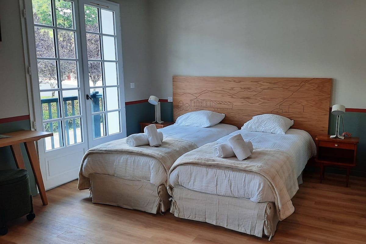 A double bed with two pillows, a bedside table, a lamp, and a view of trees through a window.