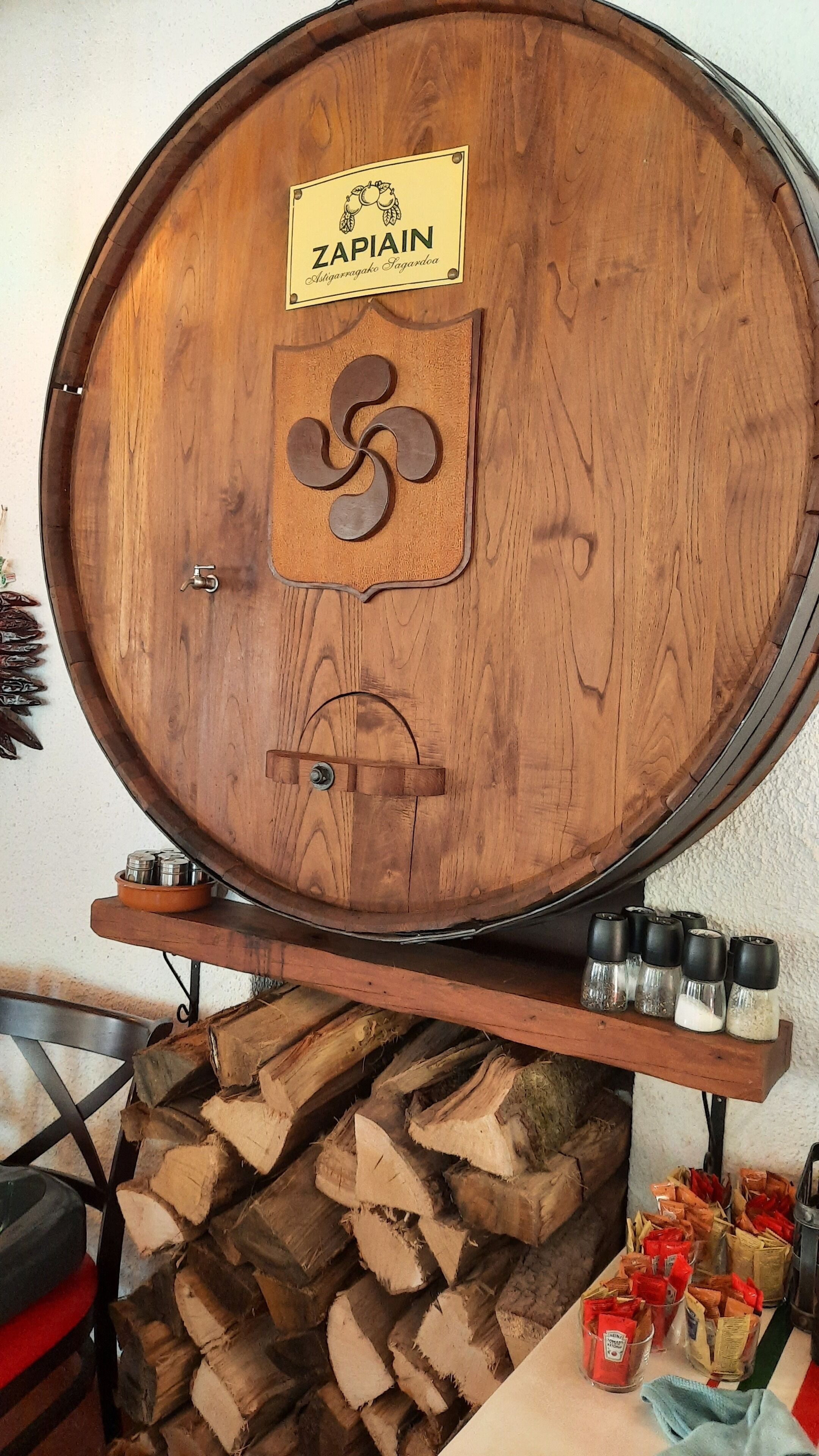 A wooden barrel with a label reading "ZAPAIN" and a logo, placed on a wooden stand with a stack of firewood and various bottles and containers nearby.