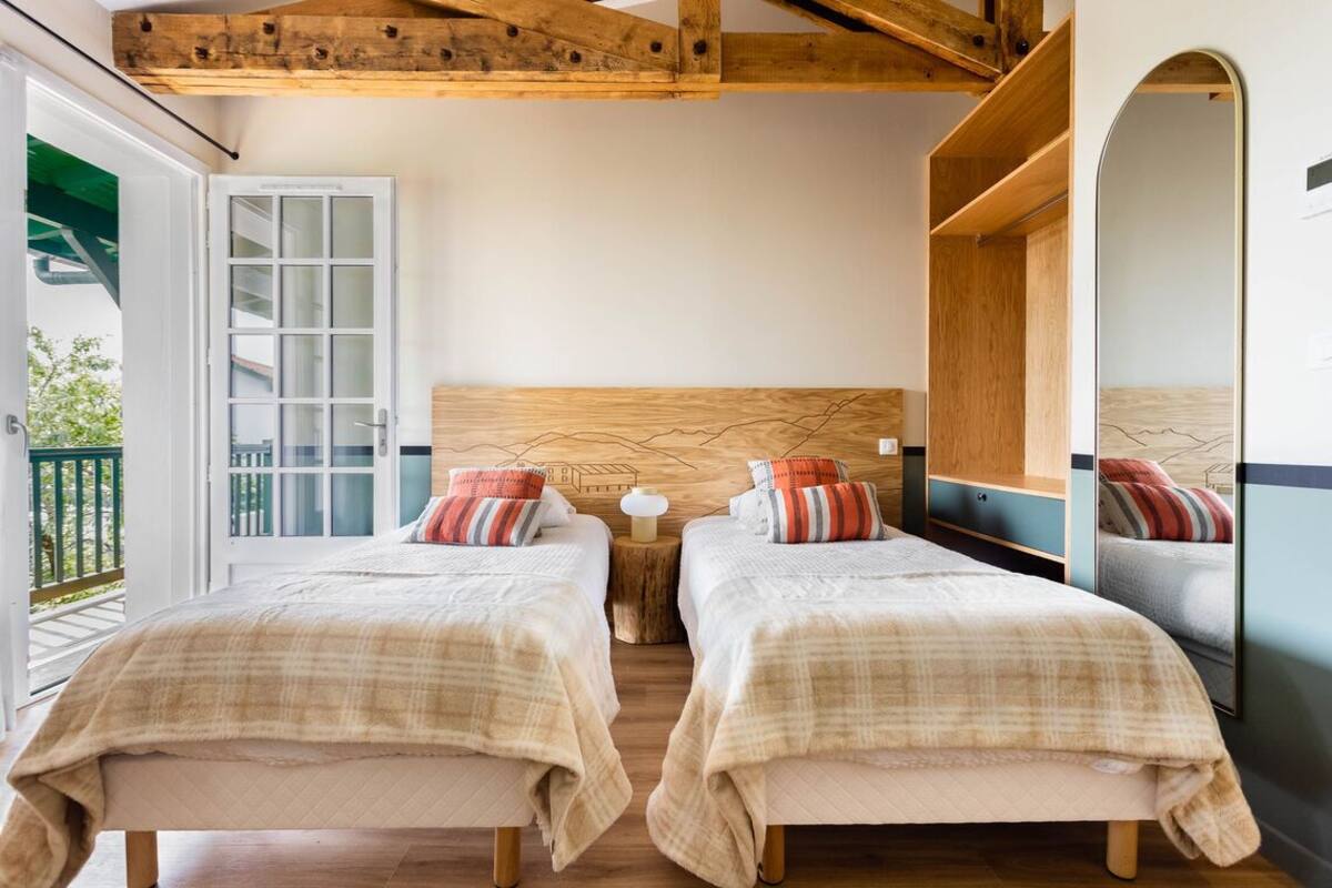 A bedroom with two beds, wooden beams, a large mirror, and a sliding glass door to a balcony.