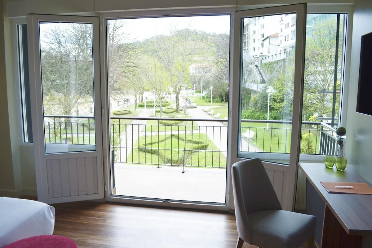 A room with a large sliding glass door leading to an outdoor balcony with a view of a landscaped garden and buildings in the background.
