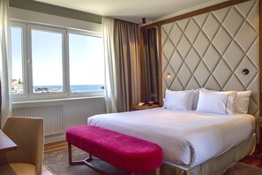 Hotelkamer met een groot bed, een bankje, een nachtkastje en uitzicht op zee door grote ramen.