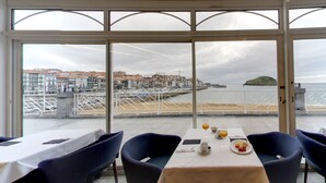 A dining area with tables and chairs, overlooking a coastal view with buildings and a pier. - Hotel Silken Palacio Uribarren (Lekeitio)