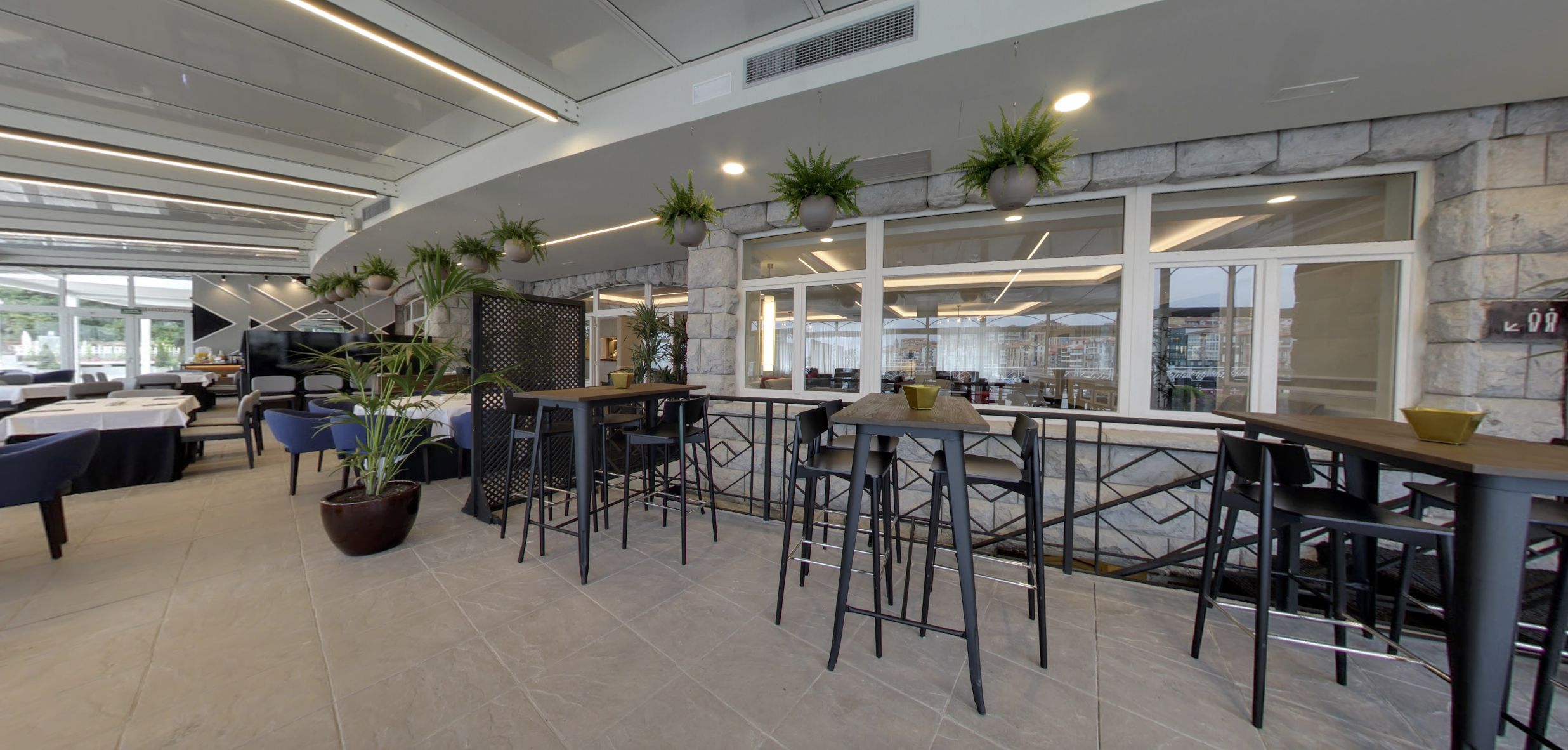 Een modern restaurant met hoge tafels en stoelen, potplanten en een stenen muur.