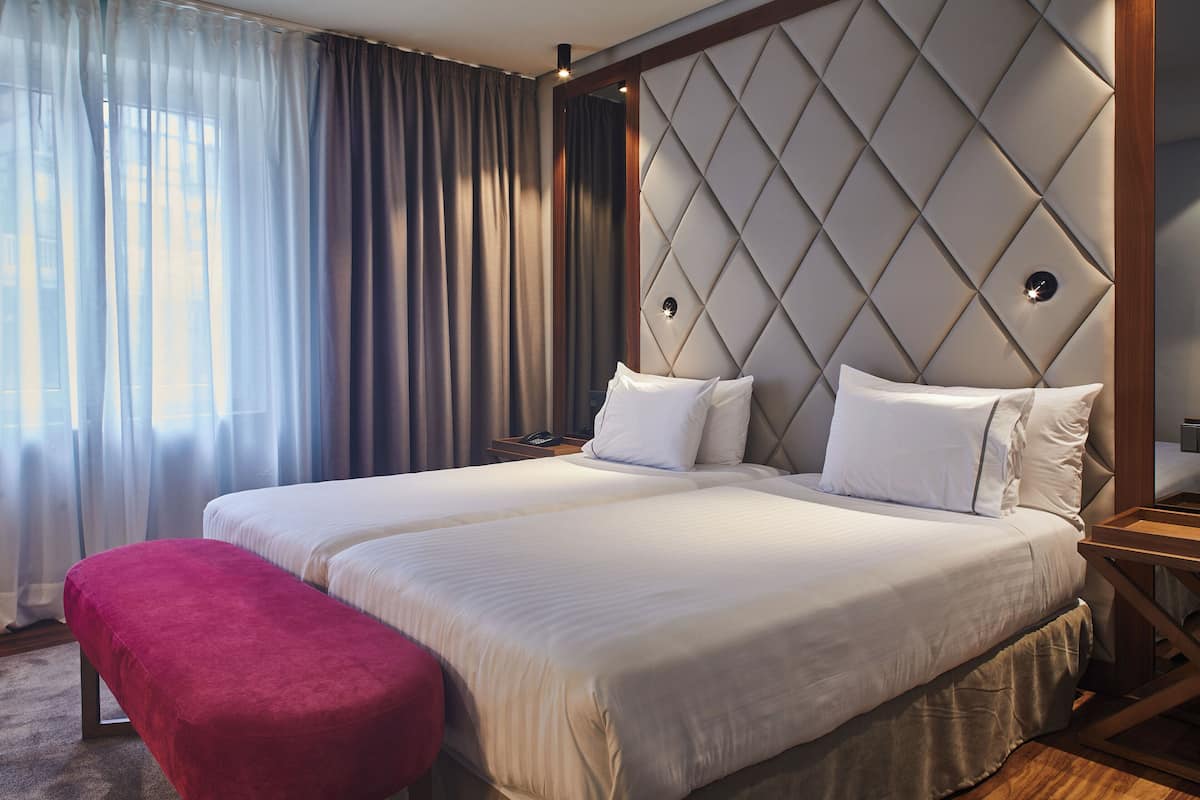 A double bed with a tufted headboard, a red bench, and a bedside table with a lamp.