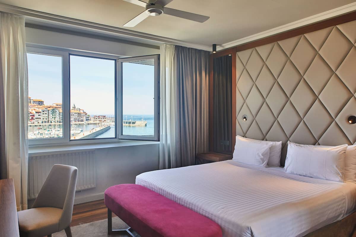 A hotel room with a large bed, a view of the cityscape through a window, a ceiling fan, and a modern headboard.
