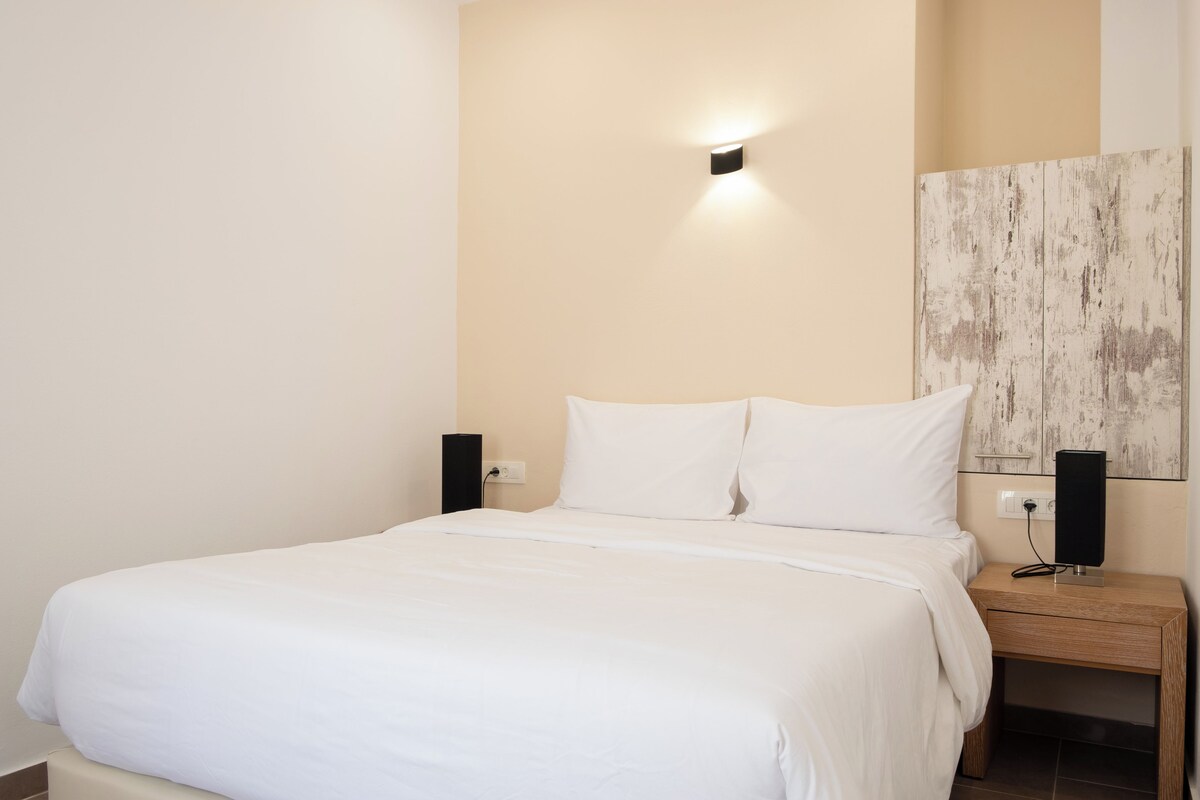 A neatly made bed with white bedding, a wall-mounted light, a wooden nightstand with a speaker, and a textured wall panel.