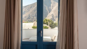 A room with a view of a mountainous landscape through a window with beige curtains. - Tristhenia Hotel (Santorini)