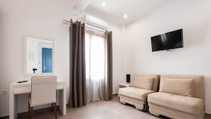 A modern room with a desk, chair, two sofas, a TV, and a window with curtains. - Tristhenia Hotel (Santorini)