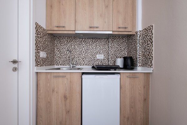 A compact kitchen with wooden cabinets, a white dishwasher, and a tiled backsplash. - Tristhenia Hotel (Santorini)