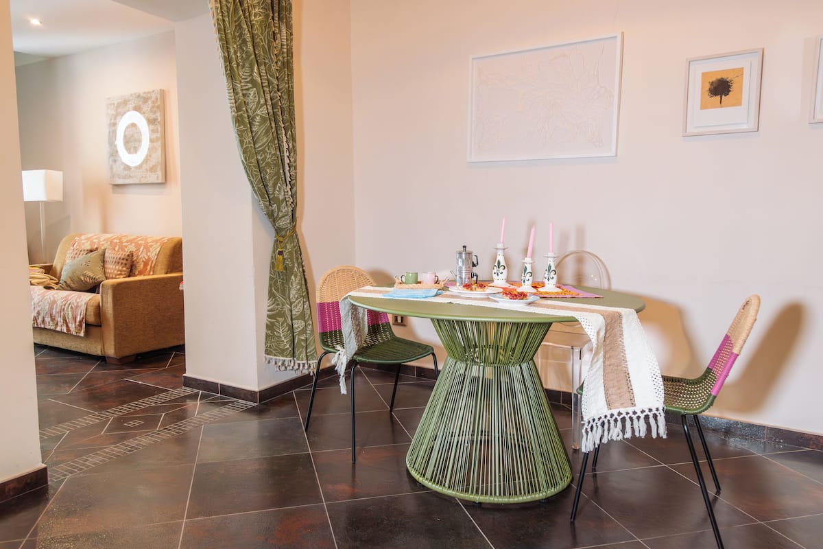 A dining area with a round table, chairs, and a tablecloth. There is a sofa in the background and a wall with framed artwork.