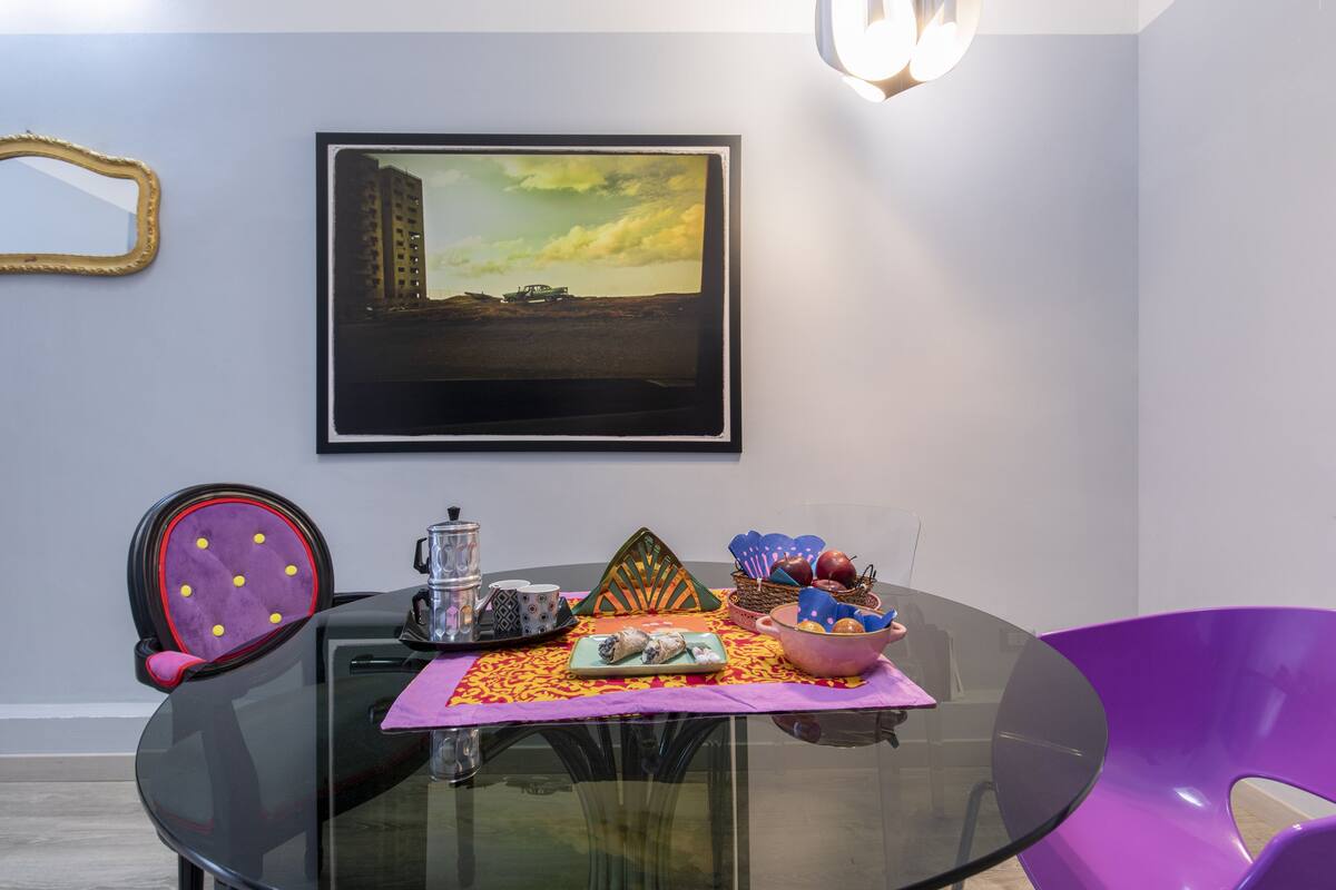 A dining area with a glass table, purple chairs, a framed picture on the wall, and a table setting with a teapot, cups, and a decorative centerpiece.
